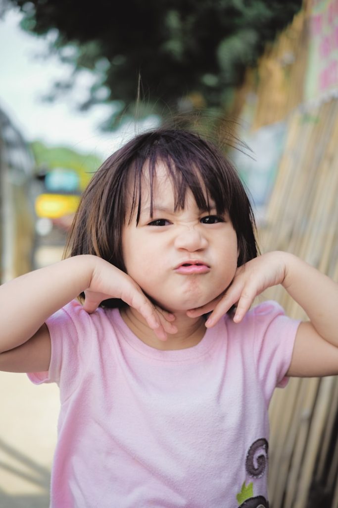 Une petite fille fait une grimace, les mains sur les joues, souriant de manière espiègle.